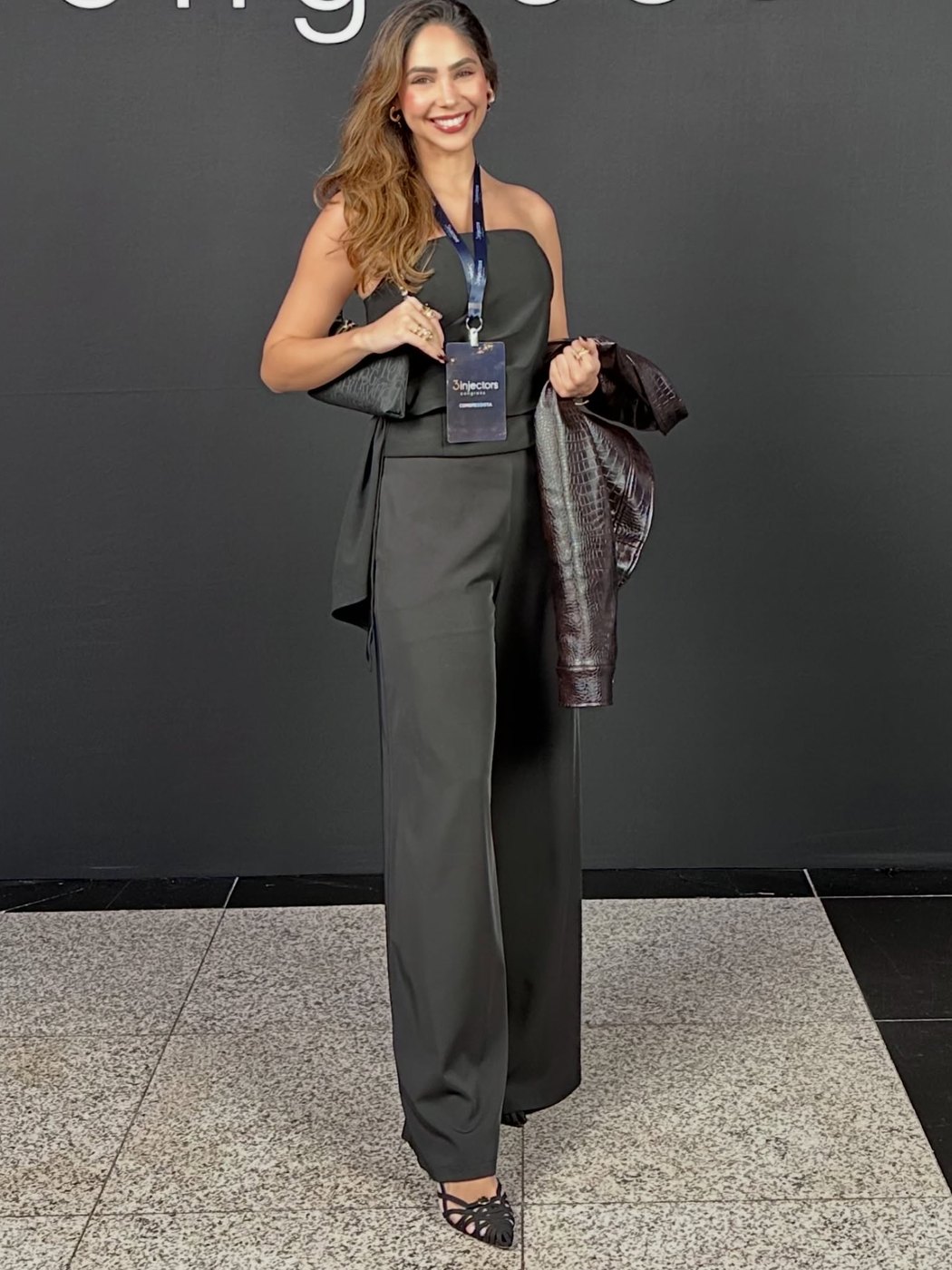 Dra. Nathália Monteiro em congresso de injetáveis, com crachá do evento e jaqueta na mão